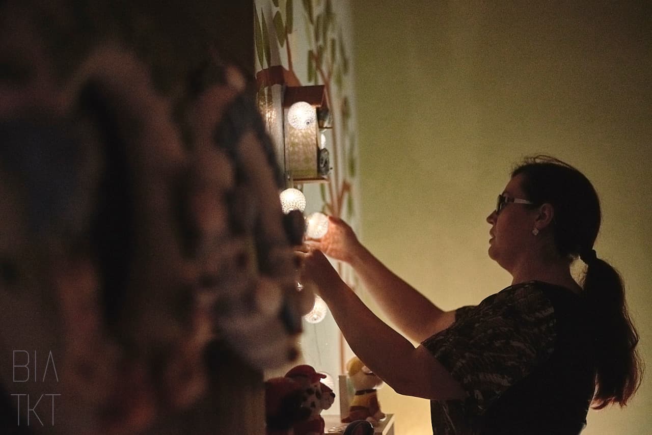 Mari Coraiola, Doula, pendura um cordão de luzes na parede de um quarto à meia-luz, preparando o ambiente para um parto.